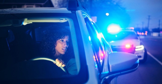 Selective focus photo of a female driver pulled over at night, a cop car with their lights on behind her.